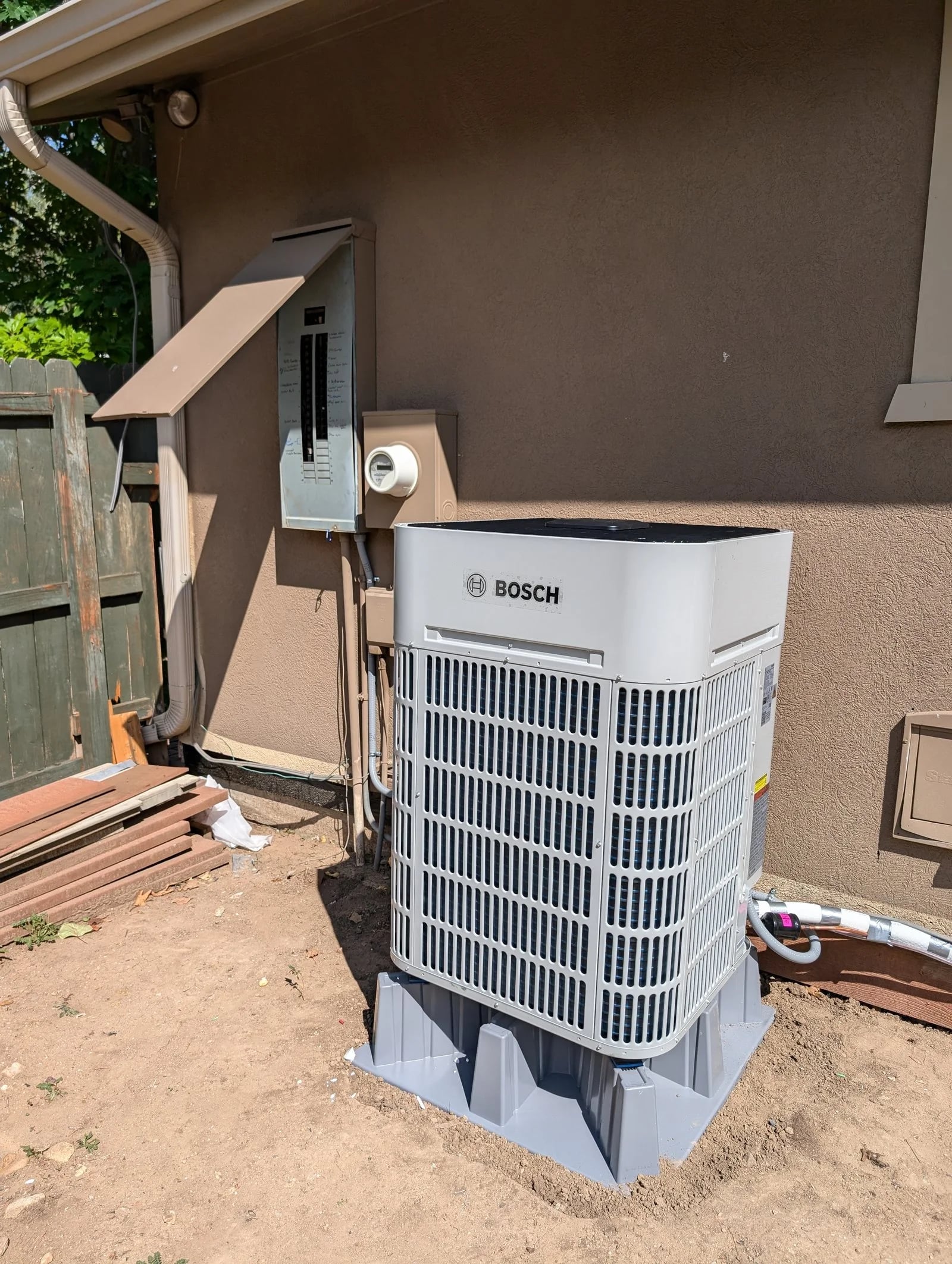 A Bosch heat pump on a composite pad beside the electrical panel on a stucco wall