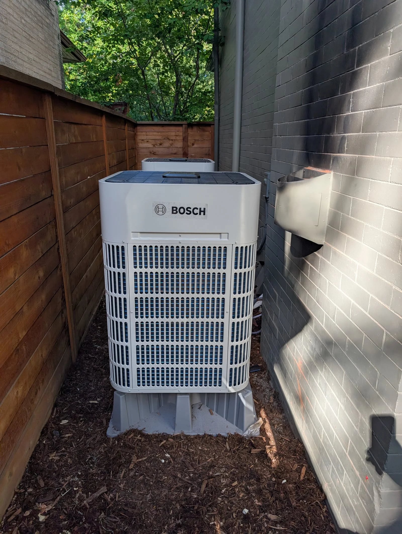 A Bosch heat pump on riser feet in a narrow side yard between the house and a wood fence