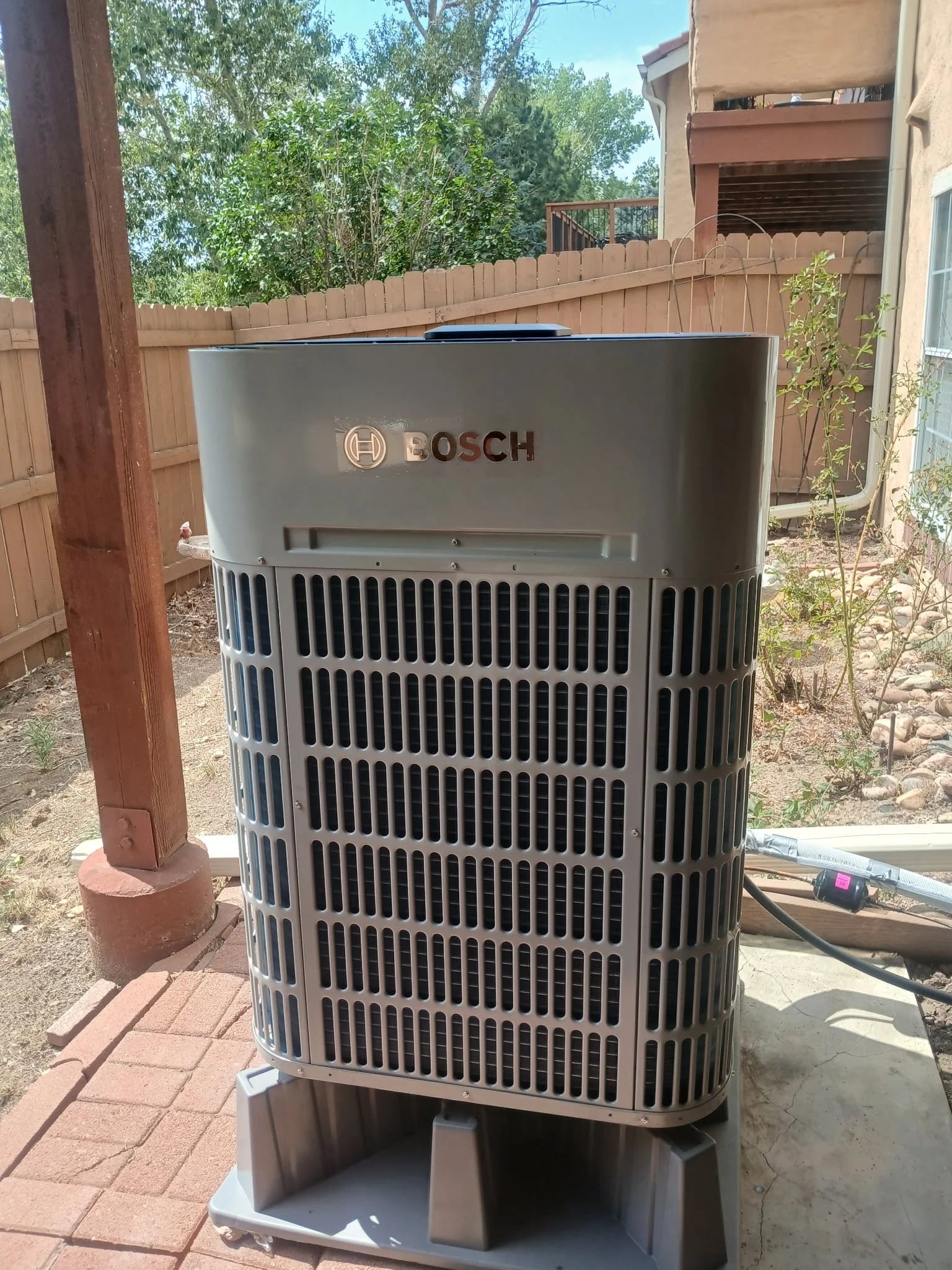 A Bosch heat pump on riser feet in a backyard, next to a wood post and fence