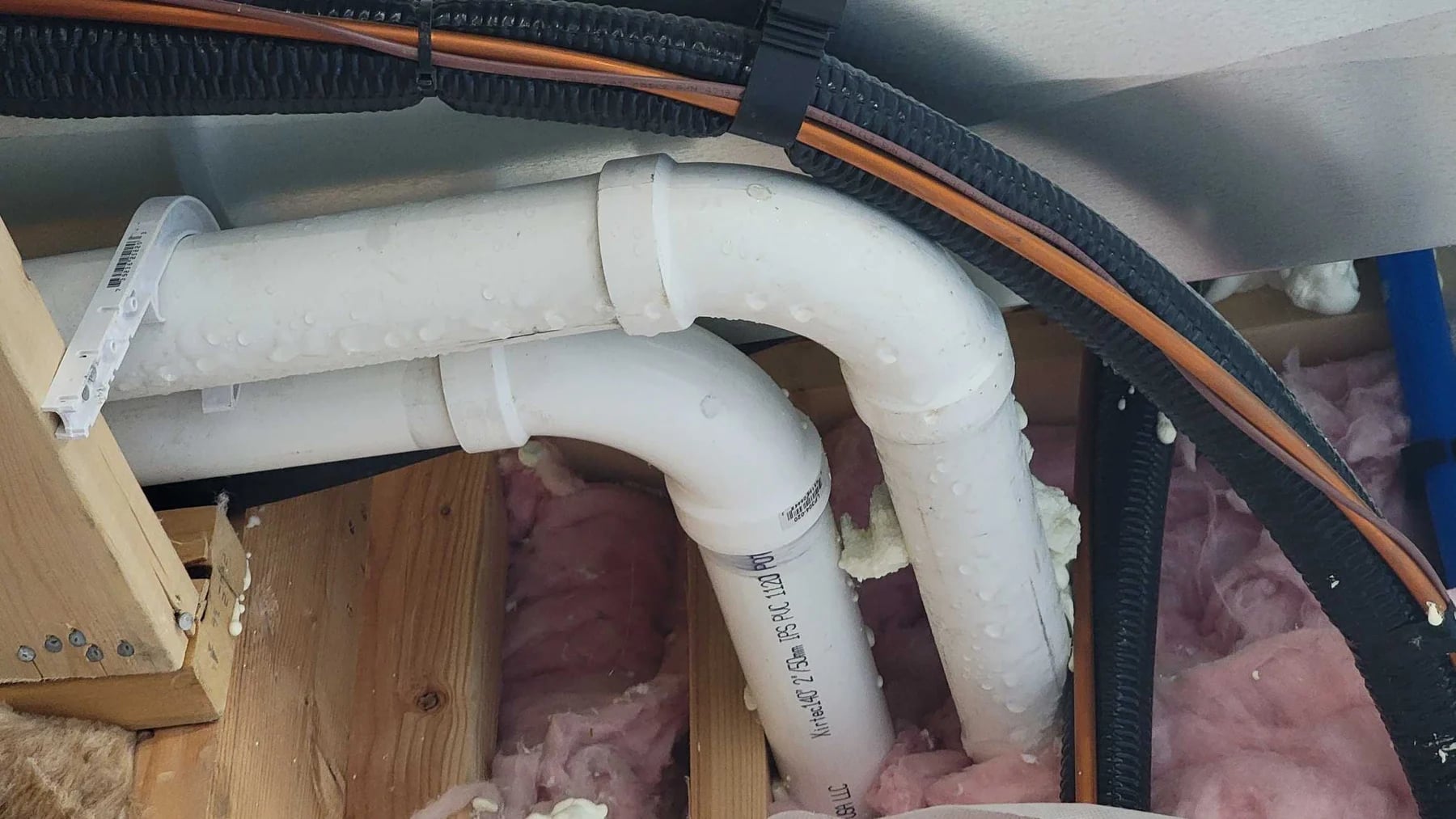 Two white PVC vent pipes crossing attic framing above pink insulation