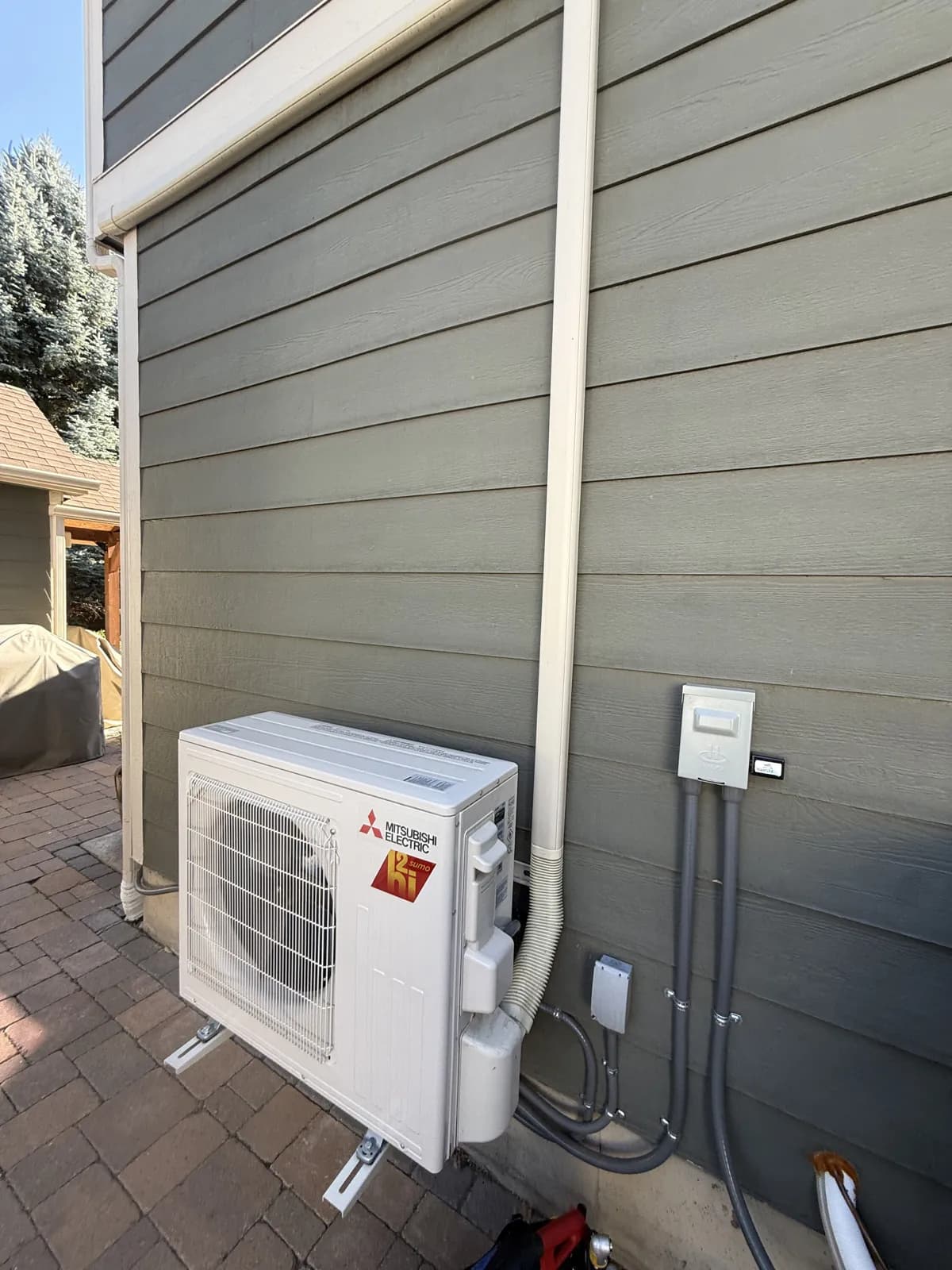 Mitsubishi compact outdoor unit on a wall stand beside gray siding with a line-set cover